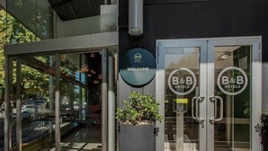 Entrada de un hotel tipo B&B con puertas de vidrio, un letrero de bienvenida y una jardinera con plantas.