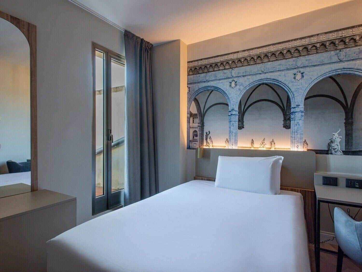 A hotel room with a bed, a desk, and a large window with curtains.