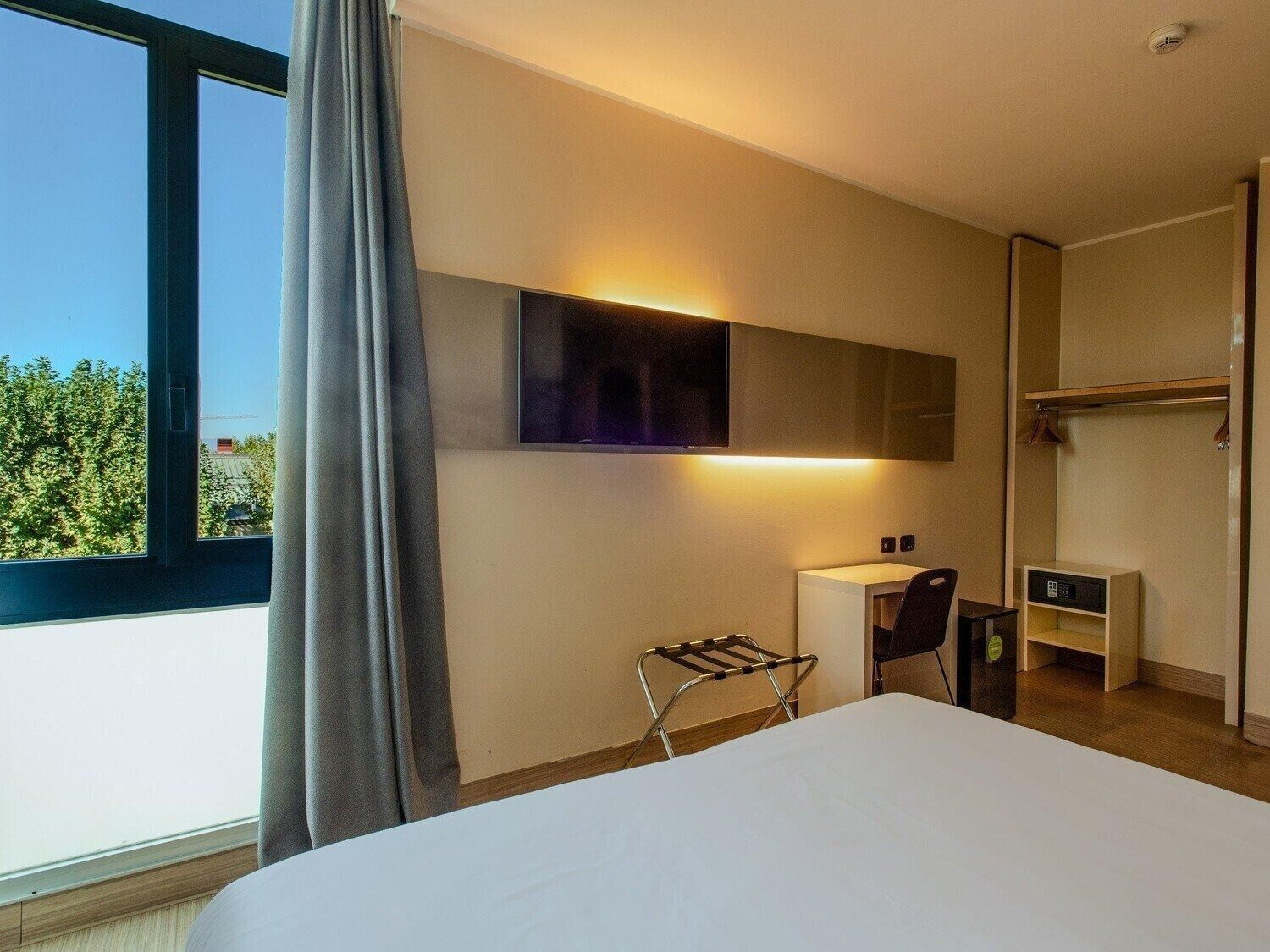 Habitación de hotel con cama, escritorio, silla, televisor montado en la pared y ventana con vista a zonas verdes.