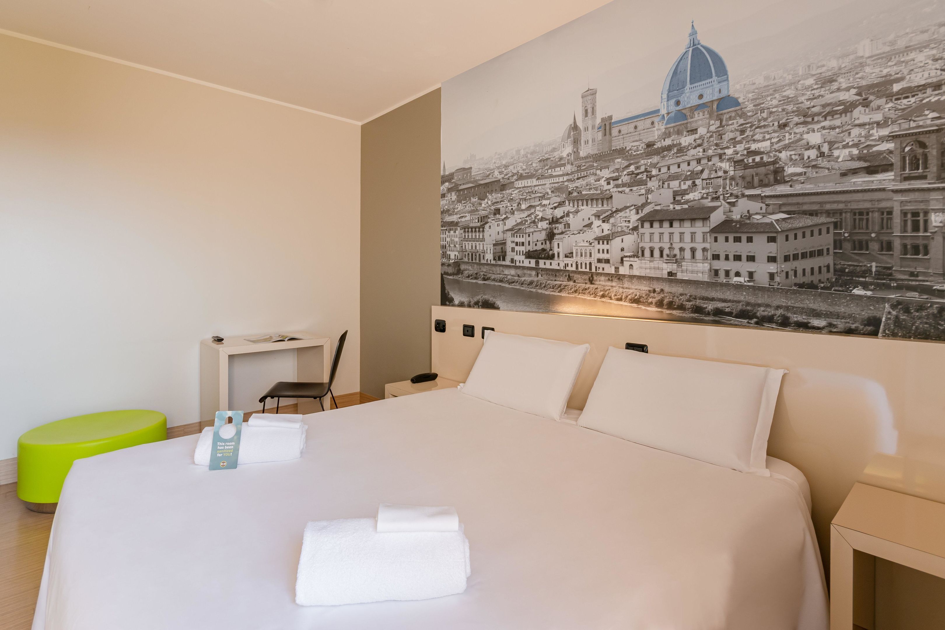 Une chambre d’hôtel avec un grand lit, un bureau, une chaise, un pouf vert et un téléviseur fixé au mur.
