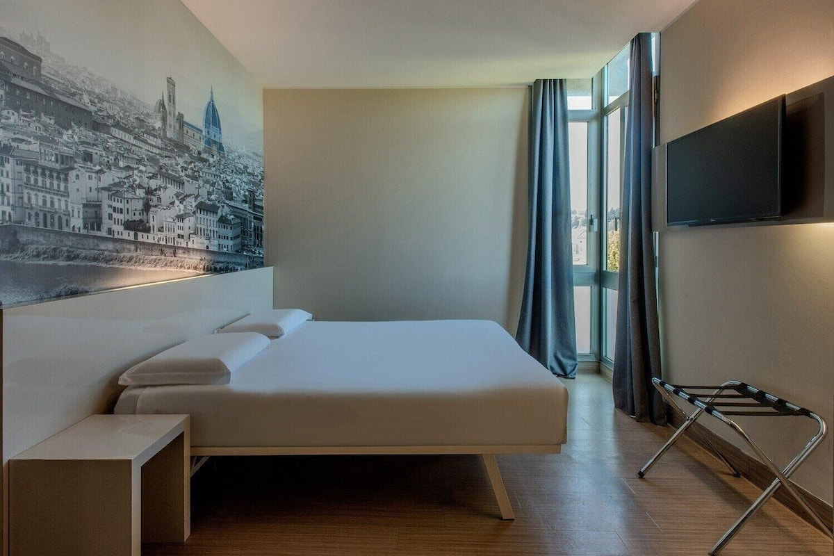 Een moderne hotelkamer met een groot bed, een flatscreen televisie, een raam met gordijnen en een stadsgezicht muurschildering.
