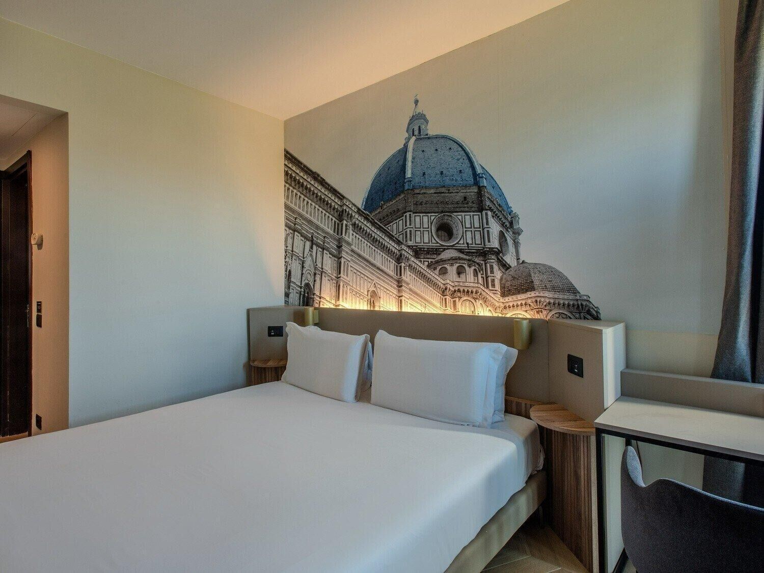 Una camera d'albergo con un letto, un comodino e un affresco raffigurante un edificio.