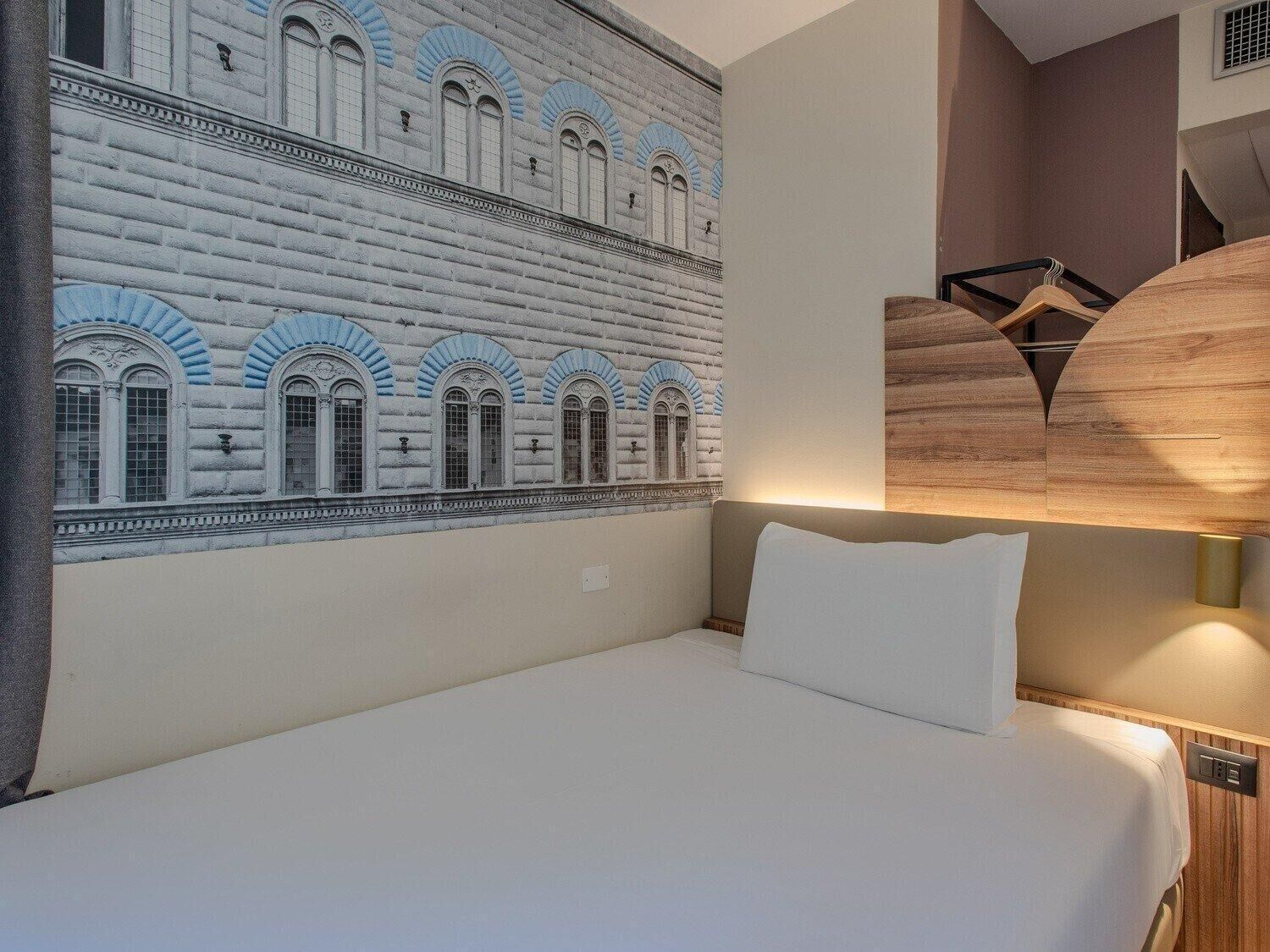 Habitación de hotel con una cama grande, cabecero de madera fijado a la pared y un mural de un edificio con ventanas de arco.