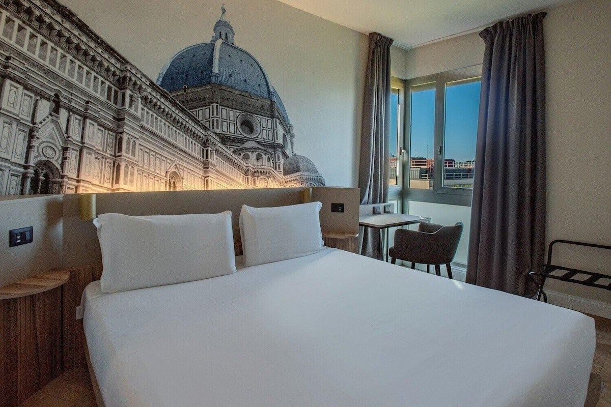 Een hotelkamer met een groot bed, een tafeltje, een stoel en uitzicht op een historisch pand.