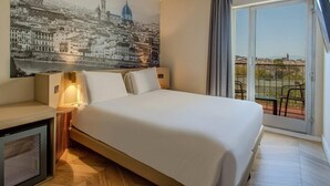 Habitación de hotel con una cama grande, una mesita de noche, una lámpara de pared, un ventanal con vistas a la ciudad y un balcón con mobiliario exterior.