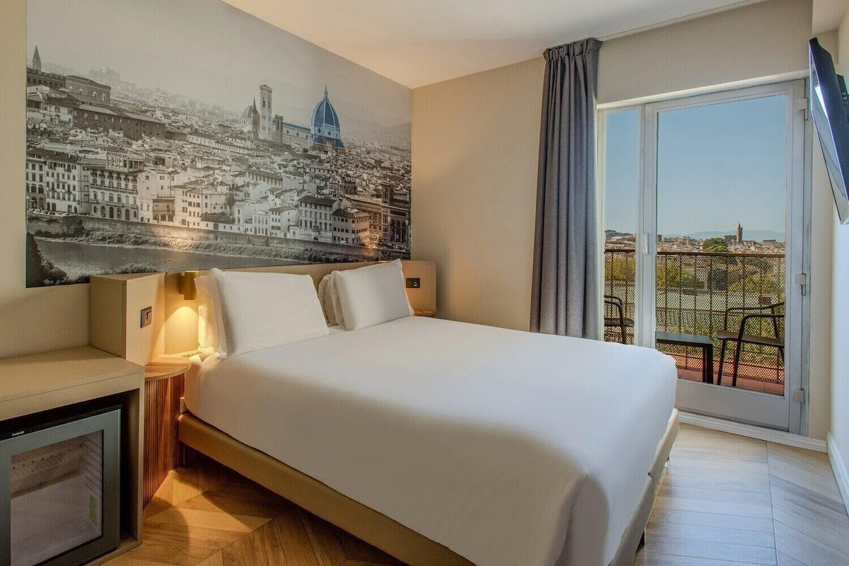 Een hotelkamer met een groot bed, een nachtkastje, een wandlamp, een groot raam met uitzicht op de stad en een balkon met buitenmeubilair.