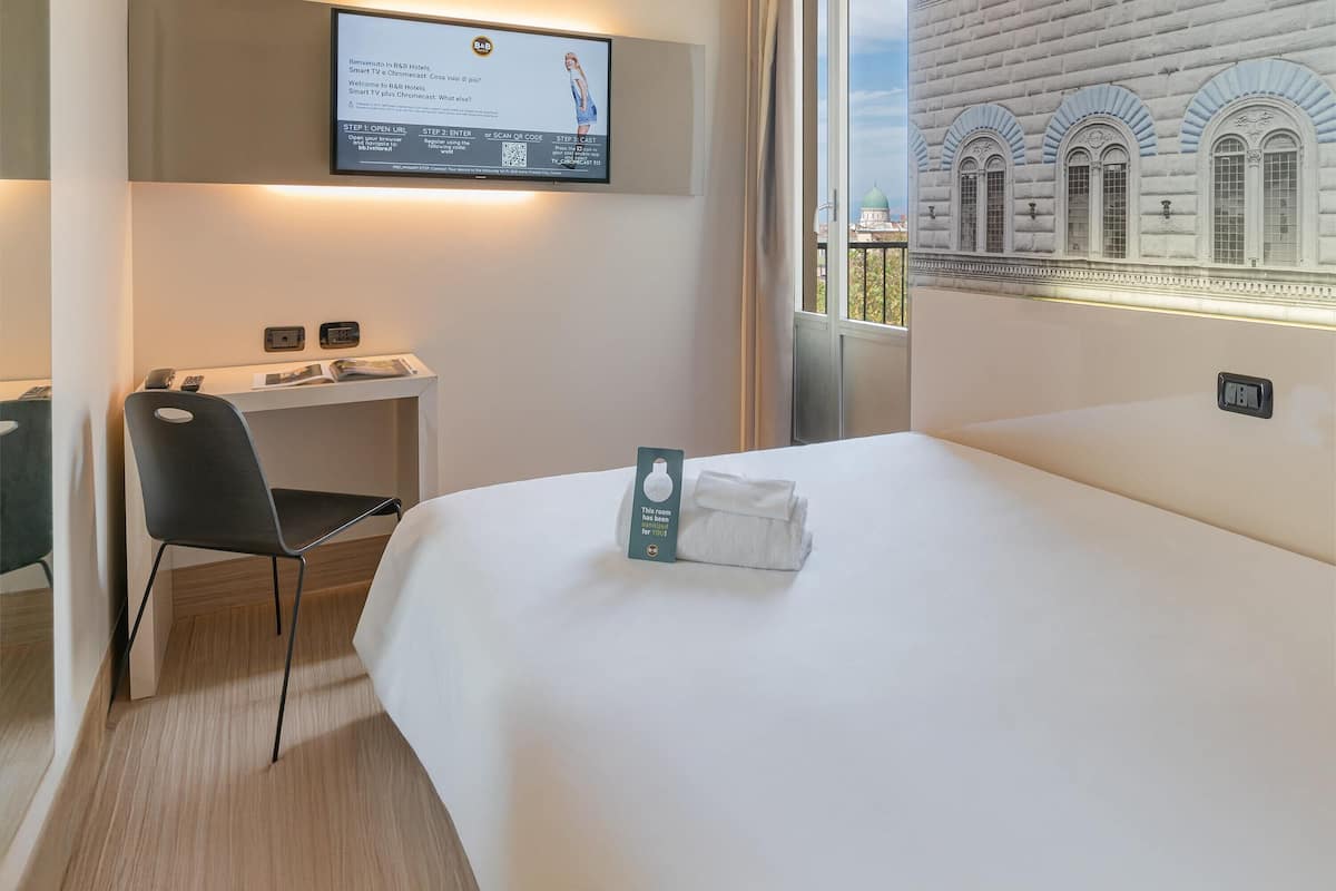 Een hotelkamer met een bed, een bureau, een stoel, een aan de muur bevestigde televisie en uitzicht op een gebouw.