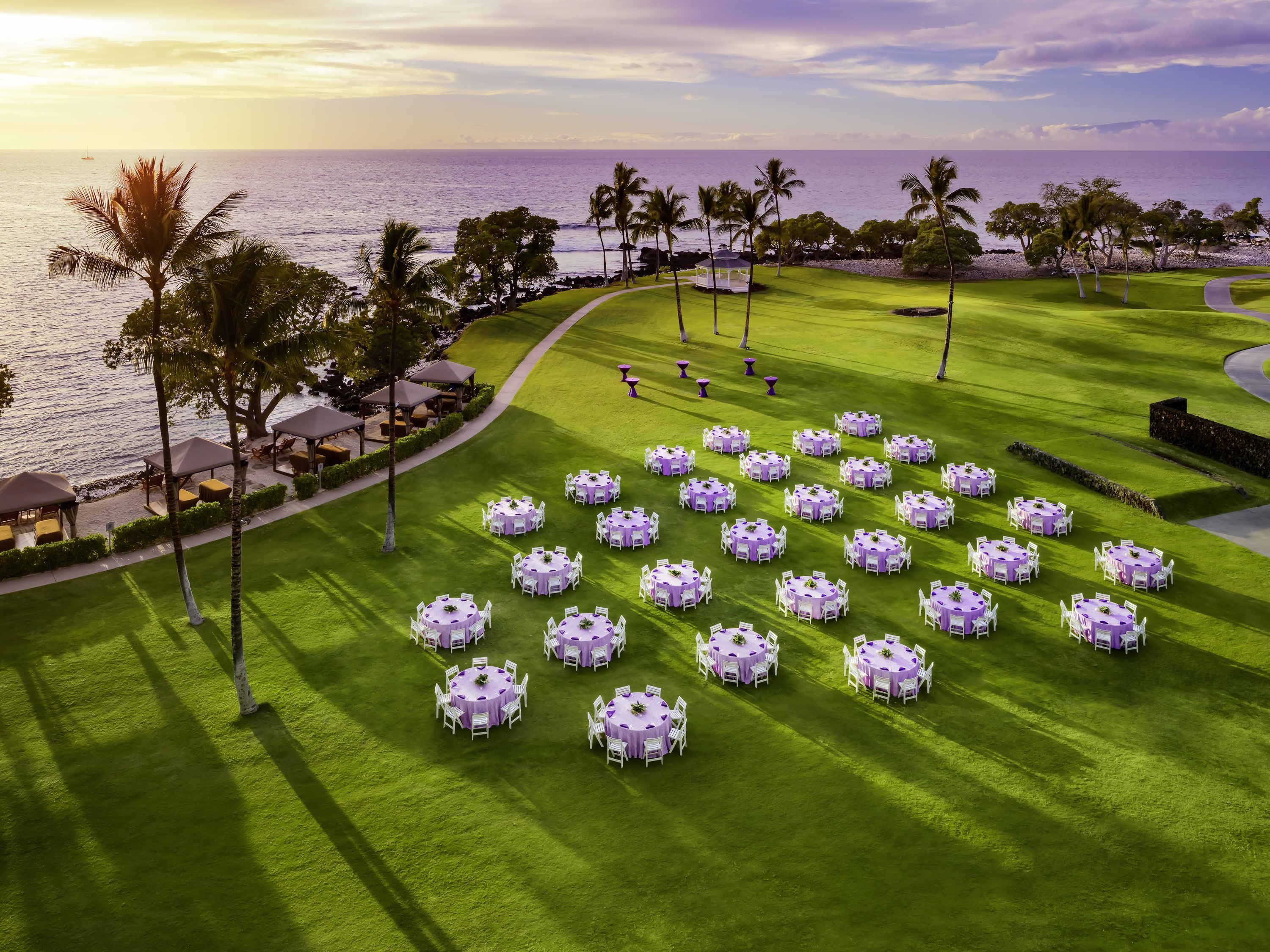 A golf course with multiple round tables set up, surrounded by palm trees and a view of the ocean.
