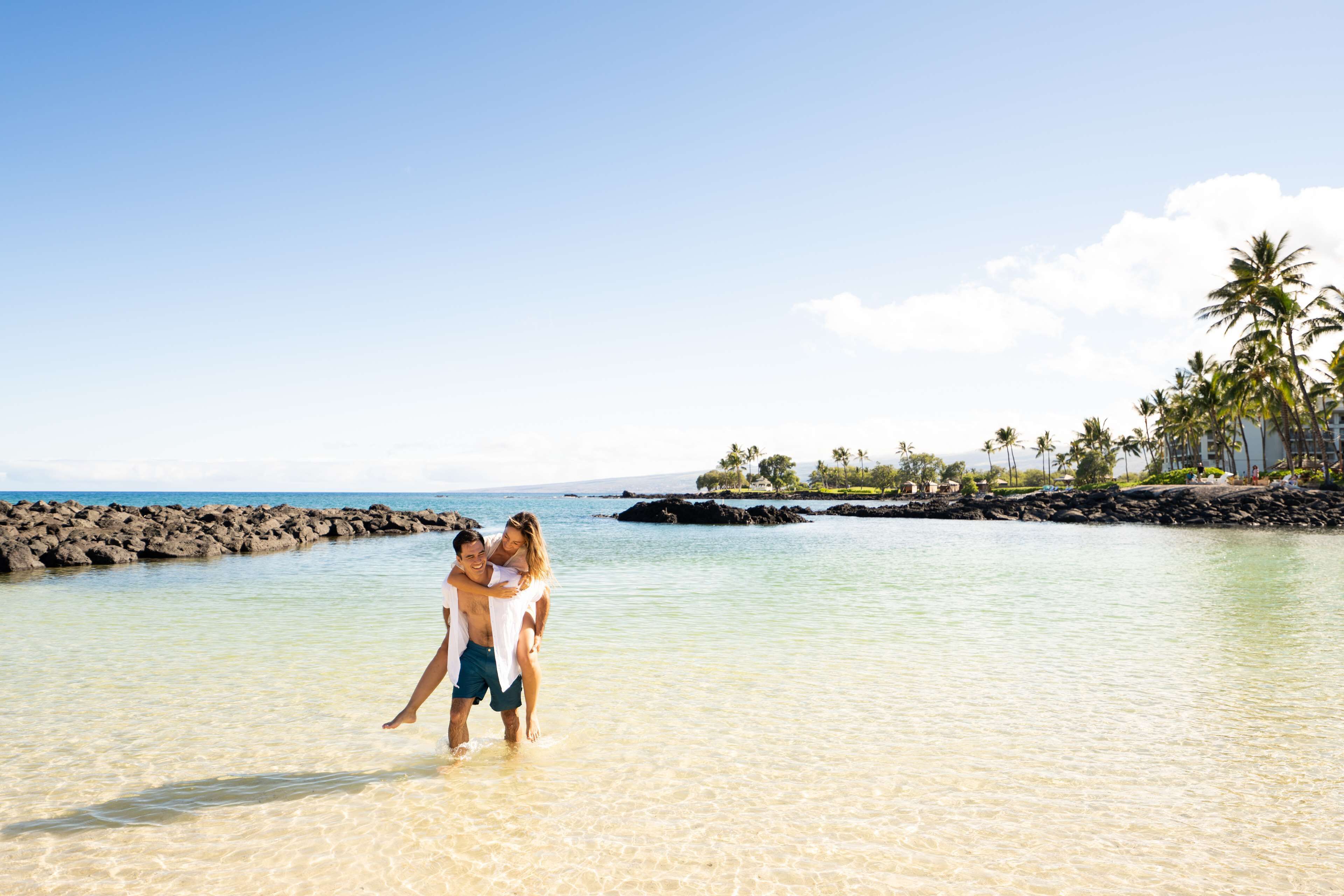 A couple embracing in shallow water near a rocky shoreline with palm trees and a clear sky.