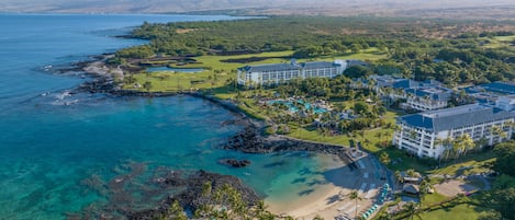 An aerial view of a resort with multiple buildings, a golf course, and a beach with clear waters.