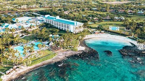 Vue aérienne d’un complexe hôtelier comprenant un grand bâtiment, une plage et une eau turquoise et cristalline.