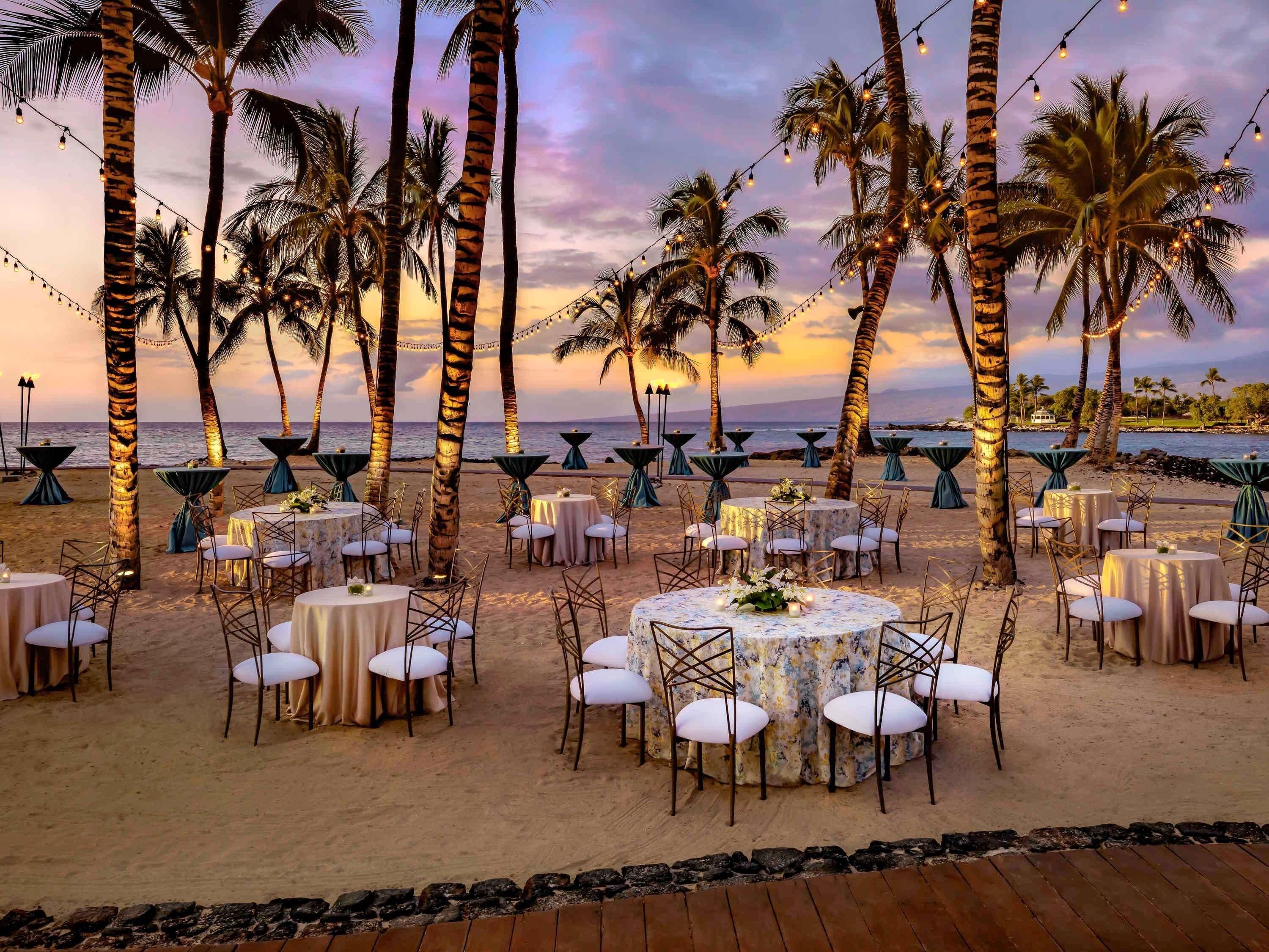 A beachside outdoor dining area with round tables and chairs, palm trees, and string lights.