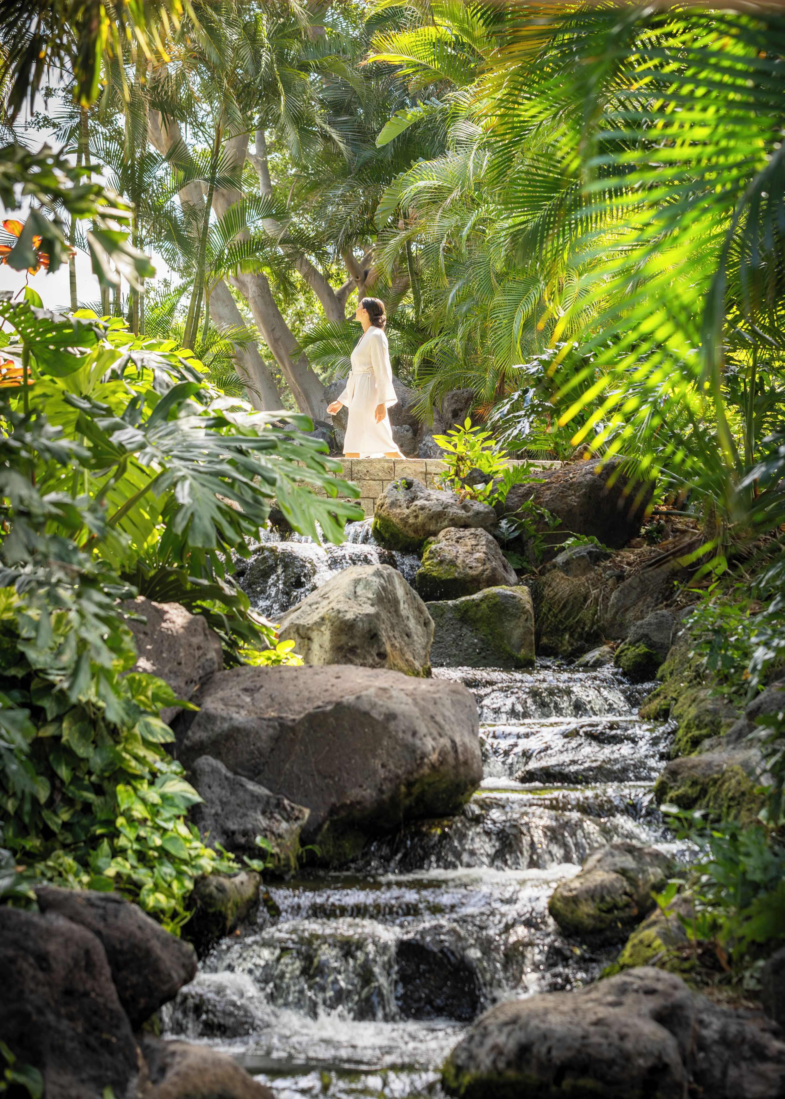 A woman in a white dress walking along a rocky stream in a lush, green garden.