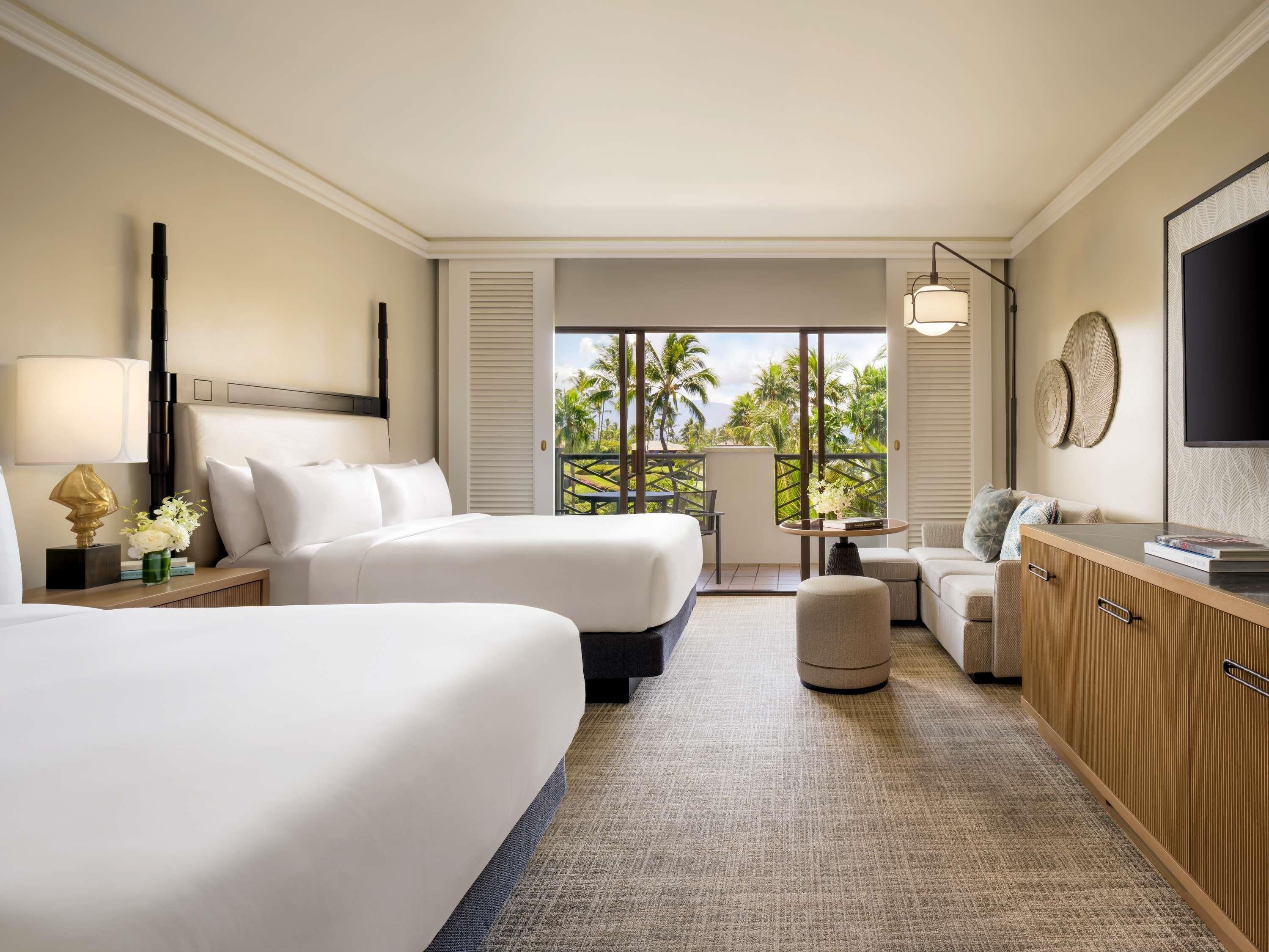 A hotel room with two beds, a sofa, a TV, and a balcony with a view of palm trees.