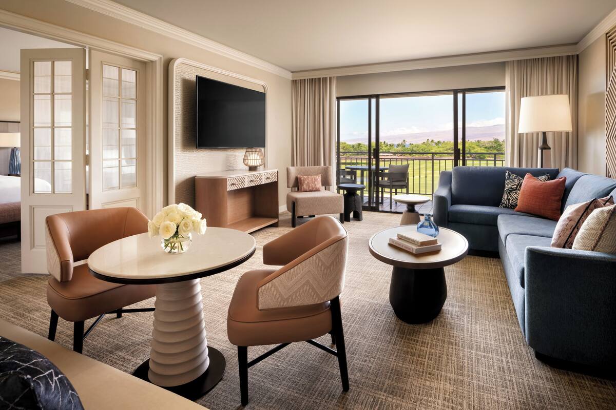 A hotel room with a balcony, a flat-screen TV, a sofa, a coffee table, and a dining area.
