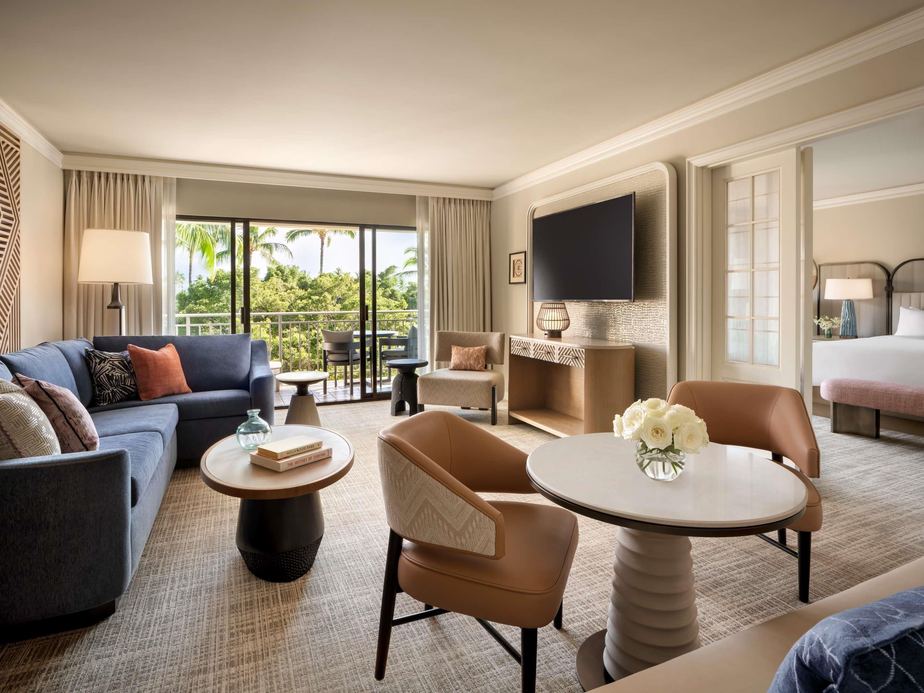 Ein Hotelzimmer mit einer Couch, Sesseln, einem kleinen Tisch, einem Fernseher und einem Balkon mit Blick auf Grünflächen.
