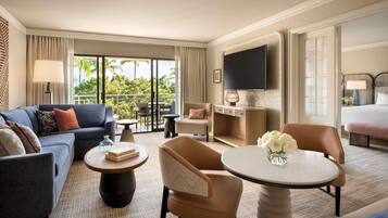 Ein Hotelzimmer mit einer Couch, Sesseln, einem kleinen Tisch, einem Fernseher und einem Balkon mit Blick auf Grünflächen.