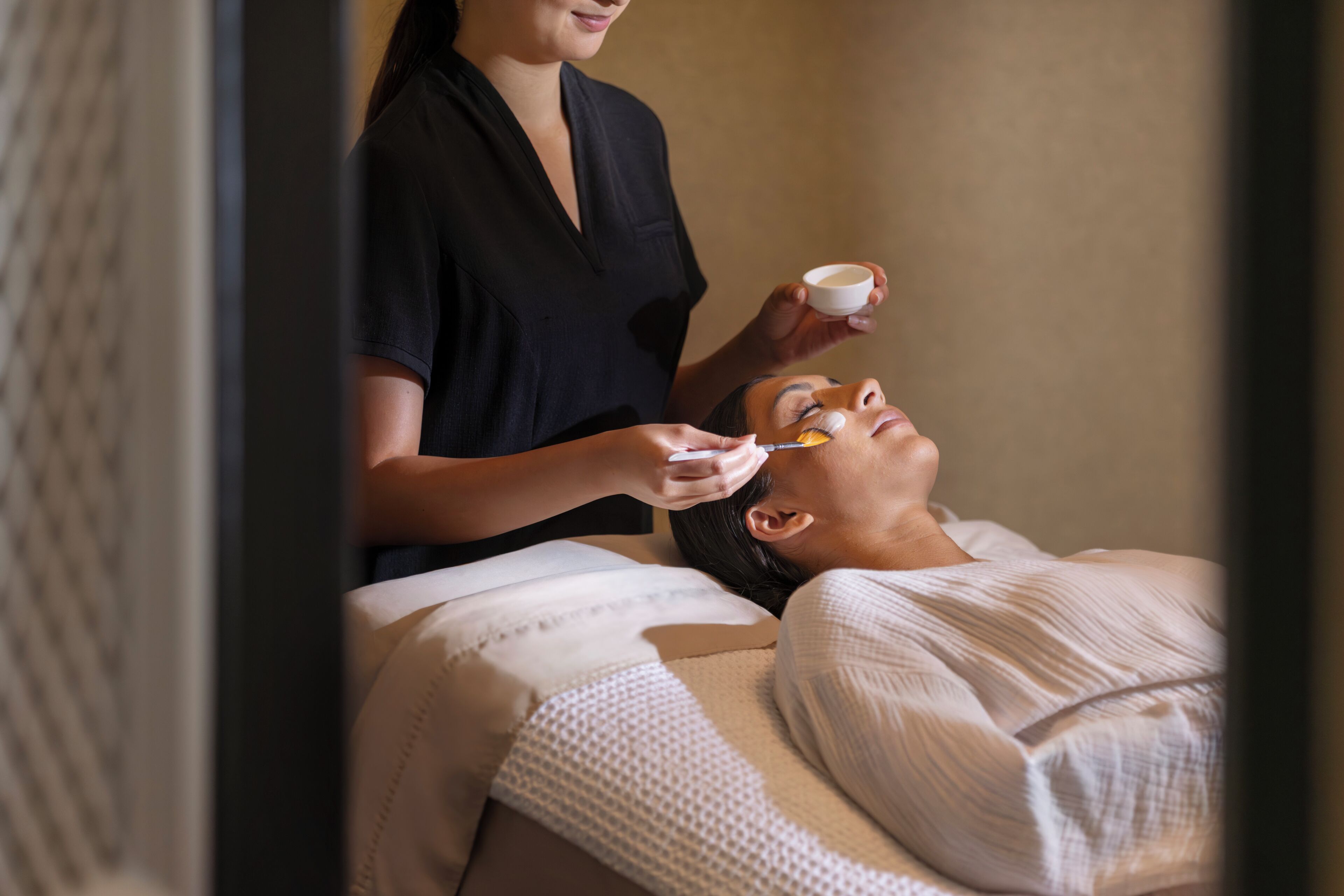 A person receiving a facial treatment in a room with a bed and a massage table.