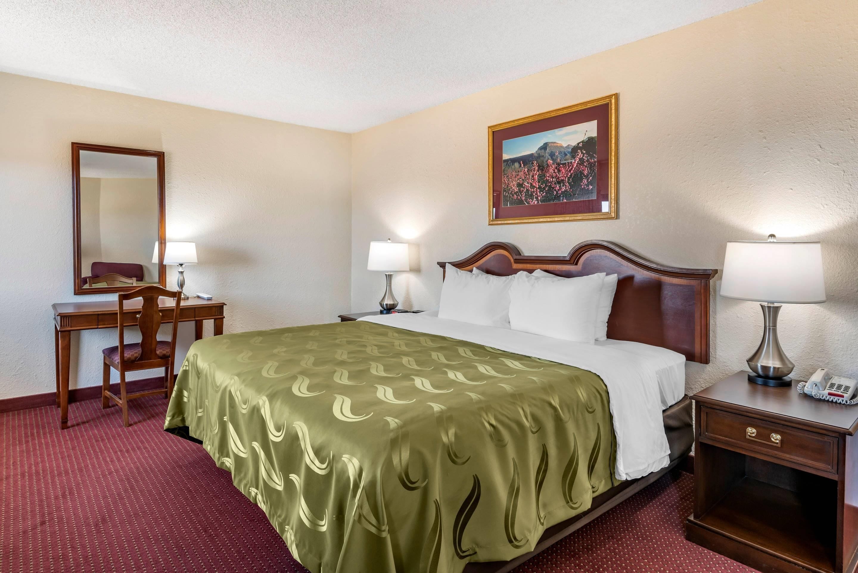 A hotel room with a bed, a desk, a chair, a nightstand, and a painting on the wall.