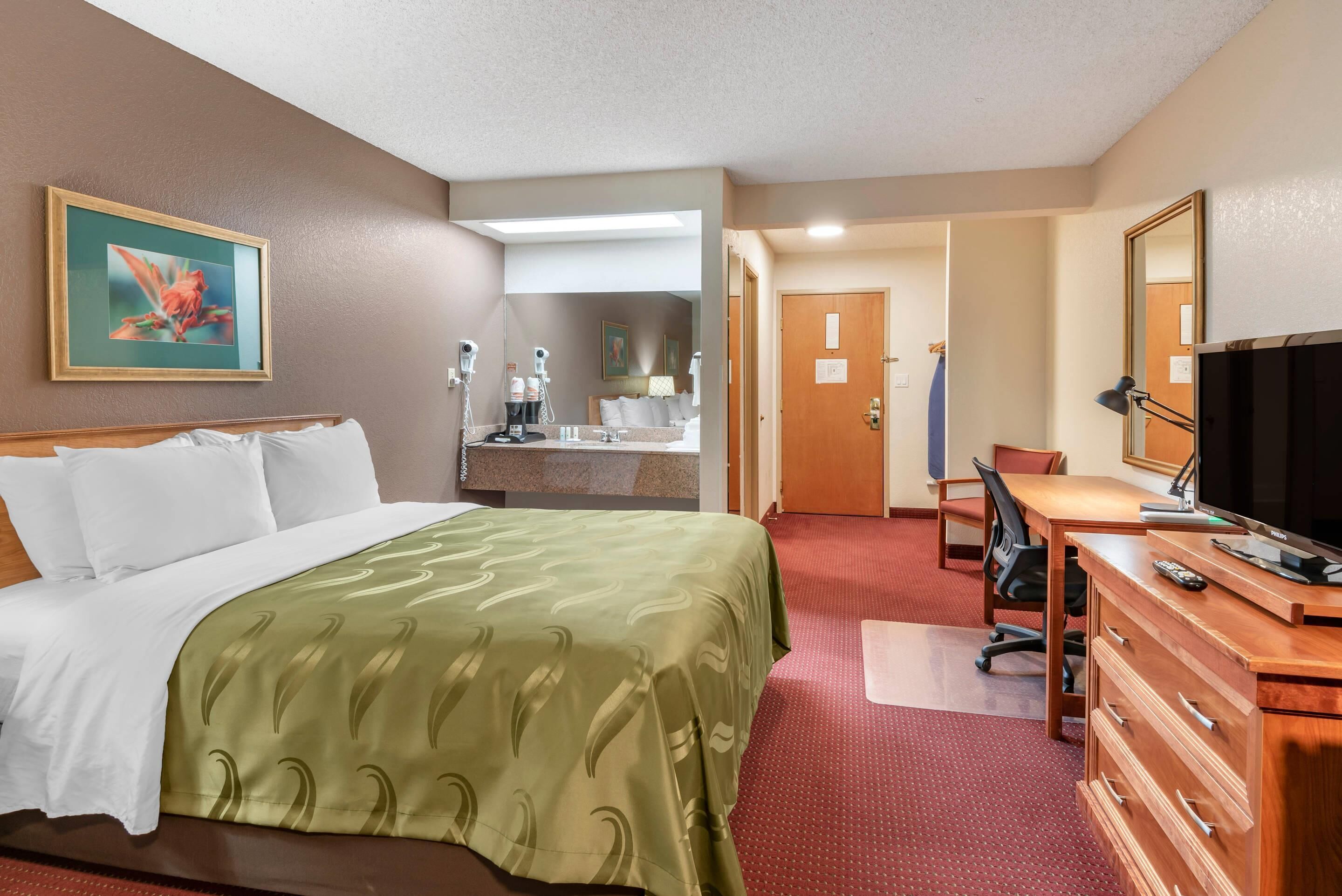 A hotel room with a bed, a desk with a computer, a TV, and a bathroom with a sink and mirror.
