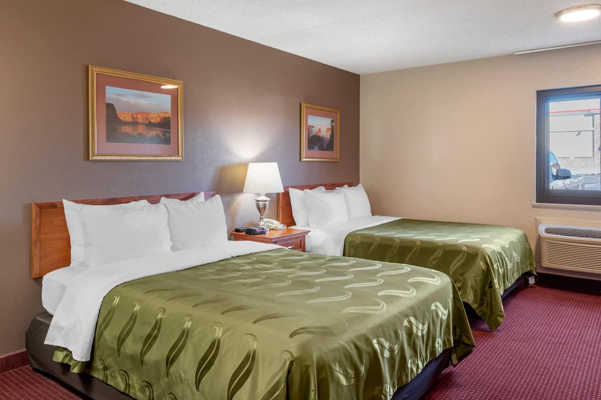 A hotel room with two beds, a nightstand with a lamp, framed pictures on the wall, and an air conditioning unit.