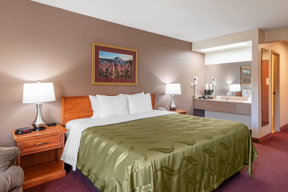 A hotel room with a large bed, two bedside tables with lamps, a small desk with a chair, a mirror, and a painting on the wall.