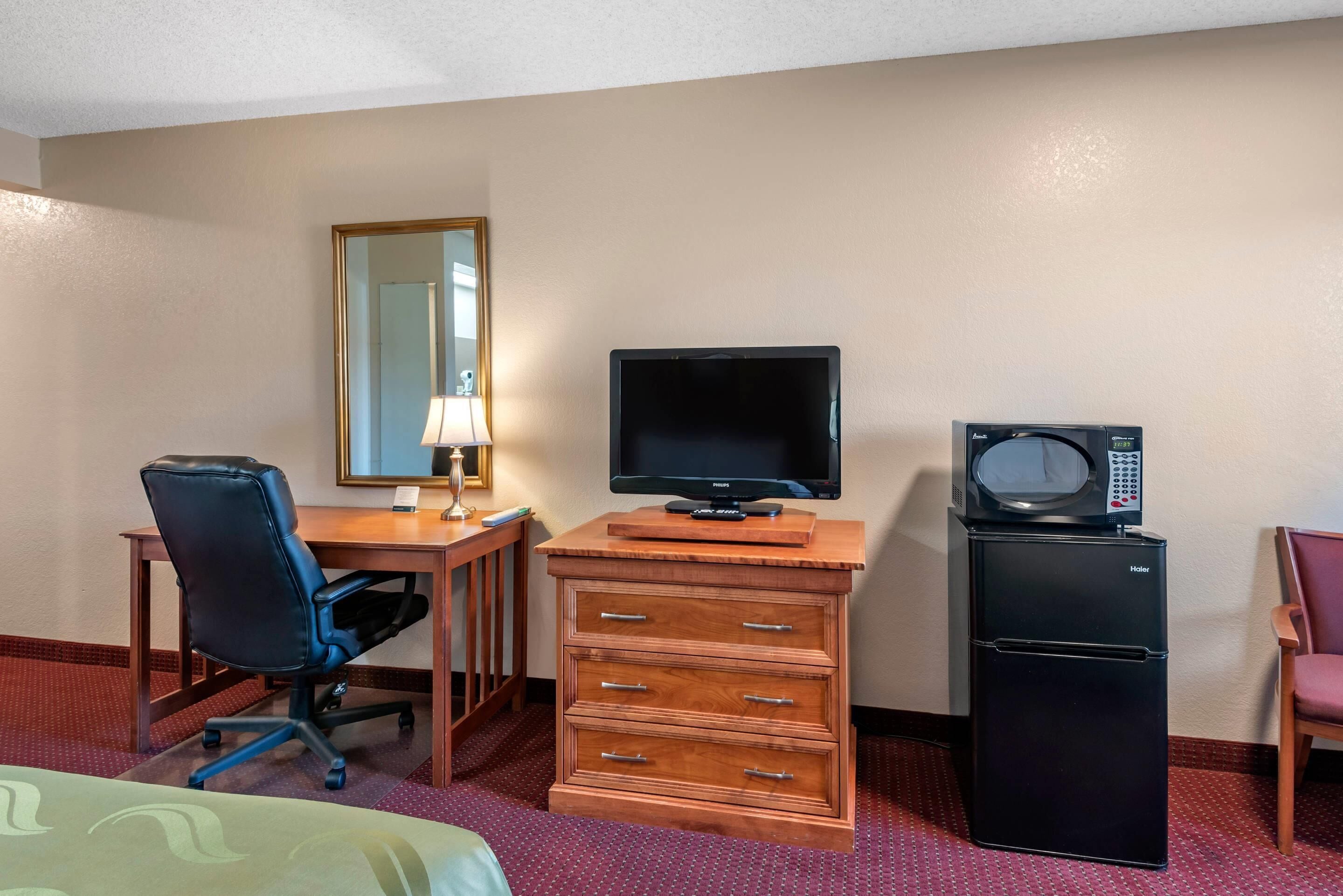 A hotel room with a wooden desk, a television, a microwave, a chair, and a dresser.
