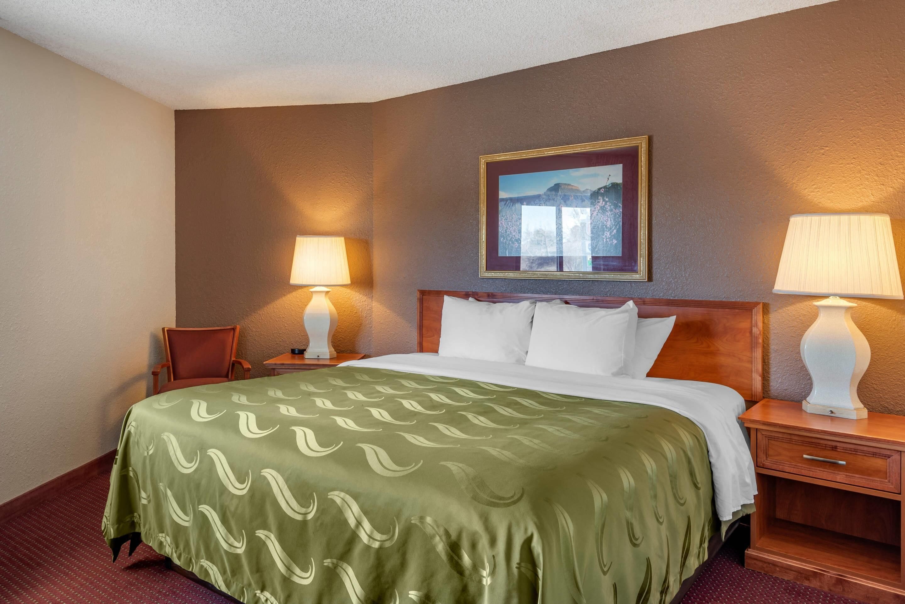 A hotel room with a large bed, two bedside lamps, a chair, and a framed picture on the wall.
