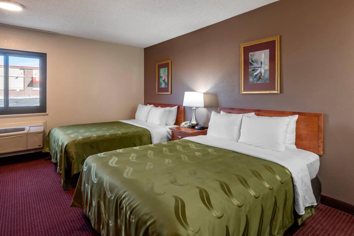 A hotel room with two beds, a window, a heater, and two framed pictures on the wall.