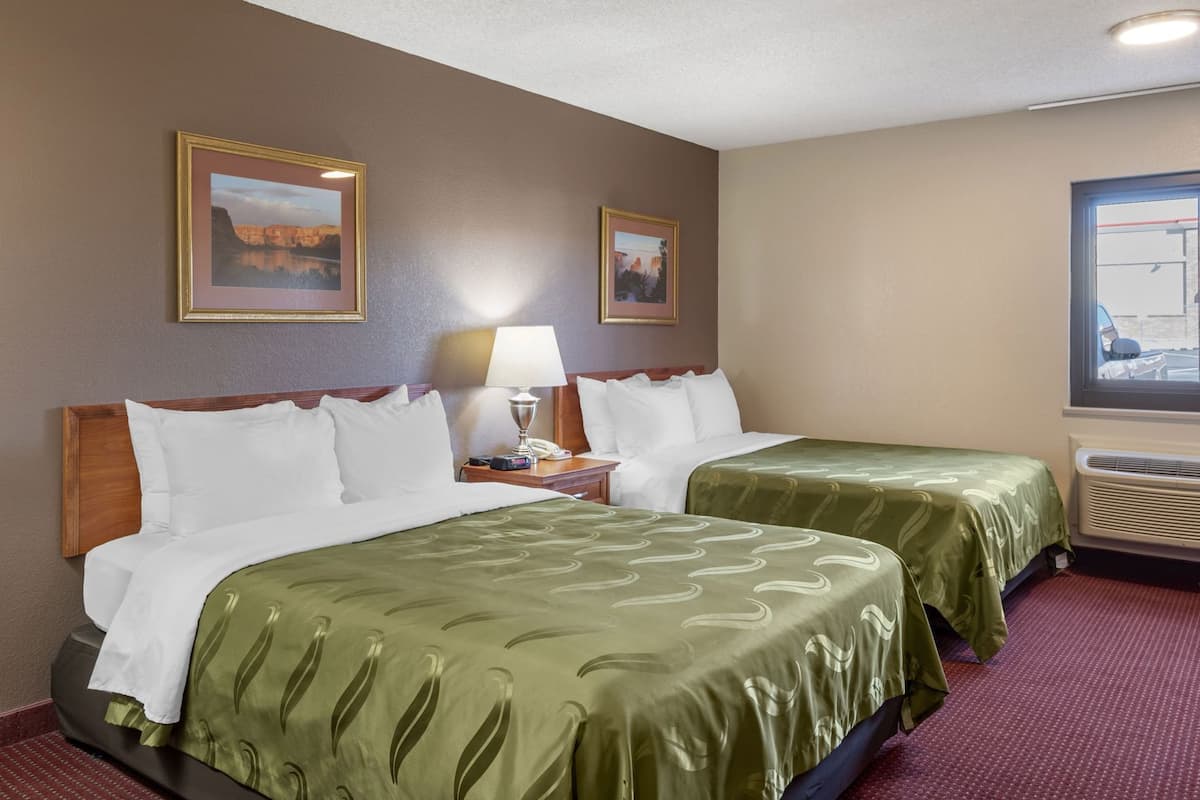 A hotel room with two beds, a nightstand, a lamp, framed pictures on the wall, and a window with a view.