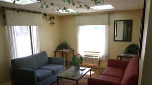 A living room with a blue sofa, a red sofa, a coffee table, and a patterned carpet. There are two windows with curtains, a potted plant on a side table, and a wall-mounted mirror. - Relax Inn (Davenport)