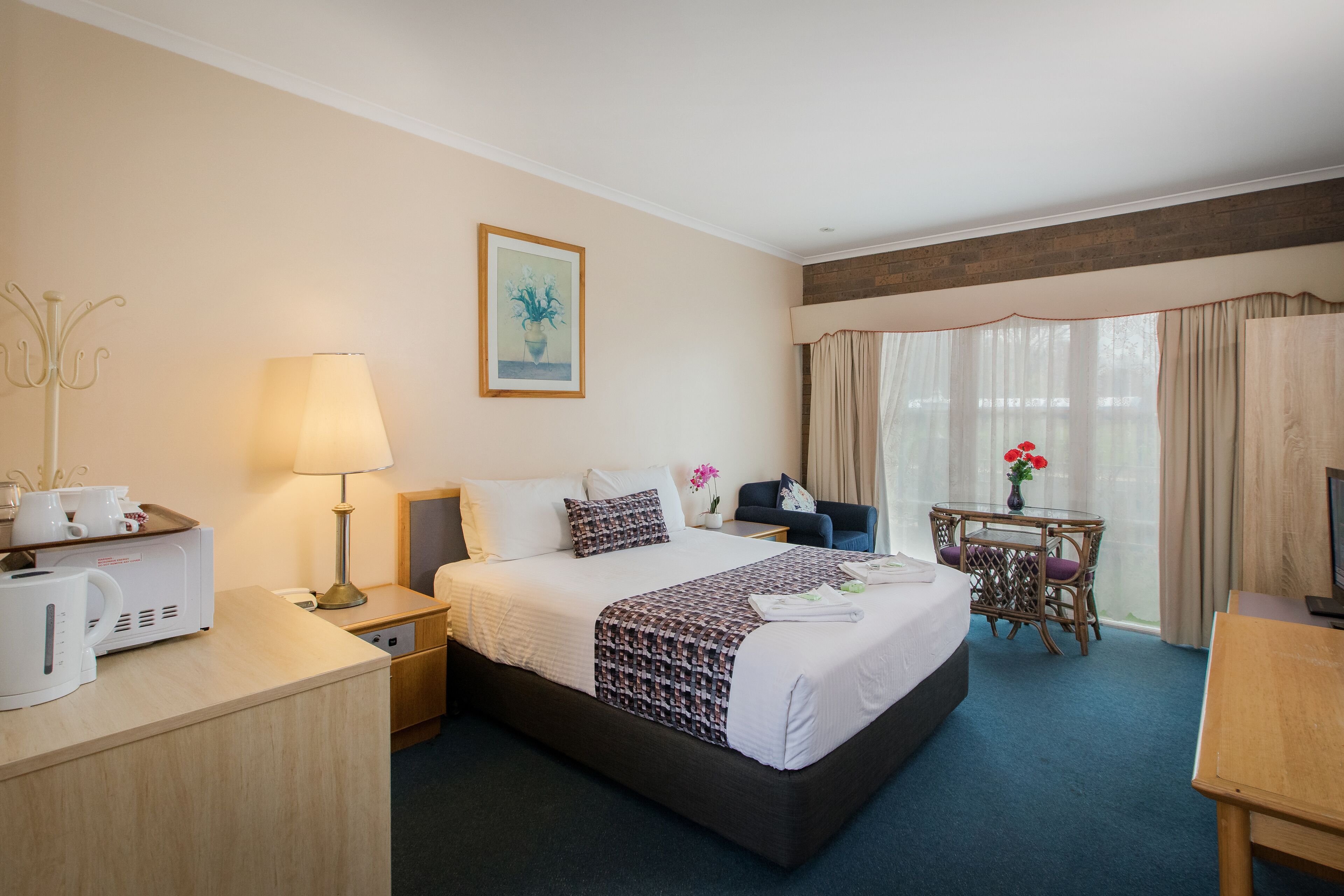 A hotel room with a bed, bedside table, lamp, coffee maker, and a small table with a vase of flowers.