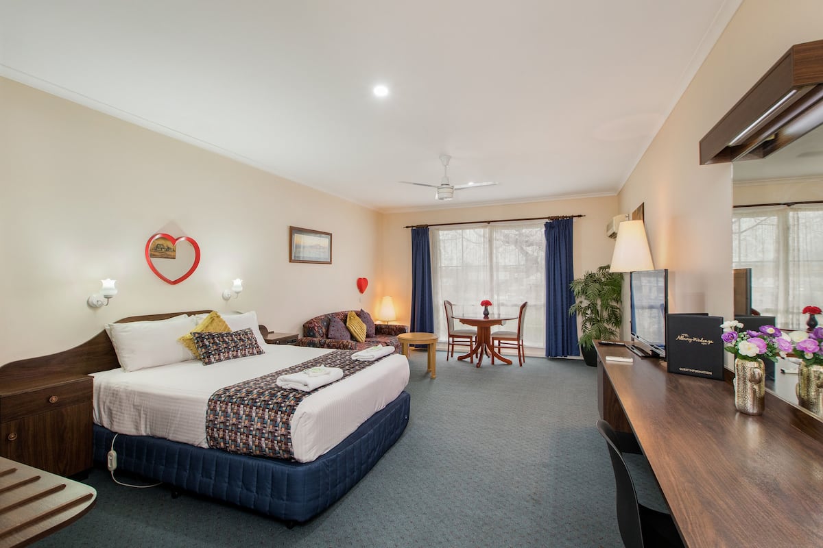 A hotel room with a large bed, a desk with a lamp, a chair, a small table, and a plant.