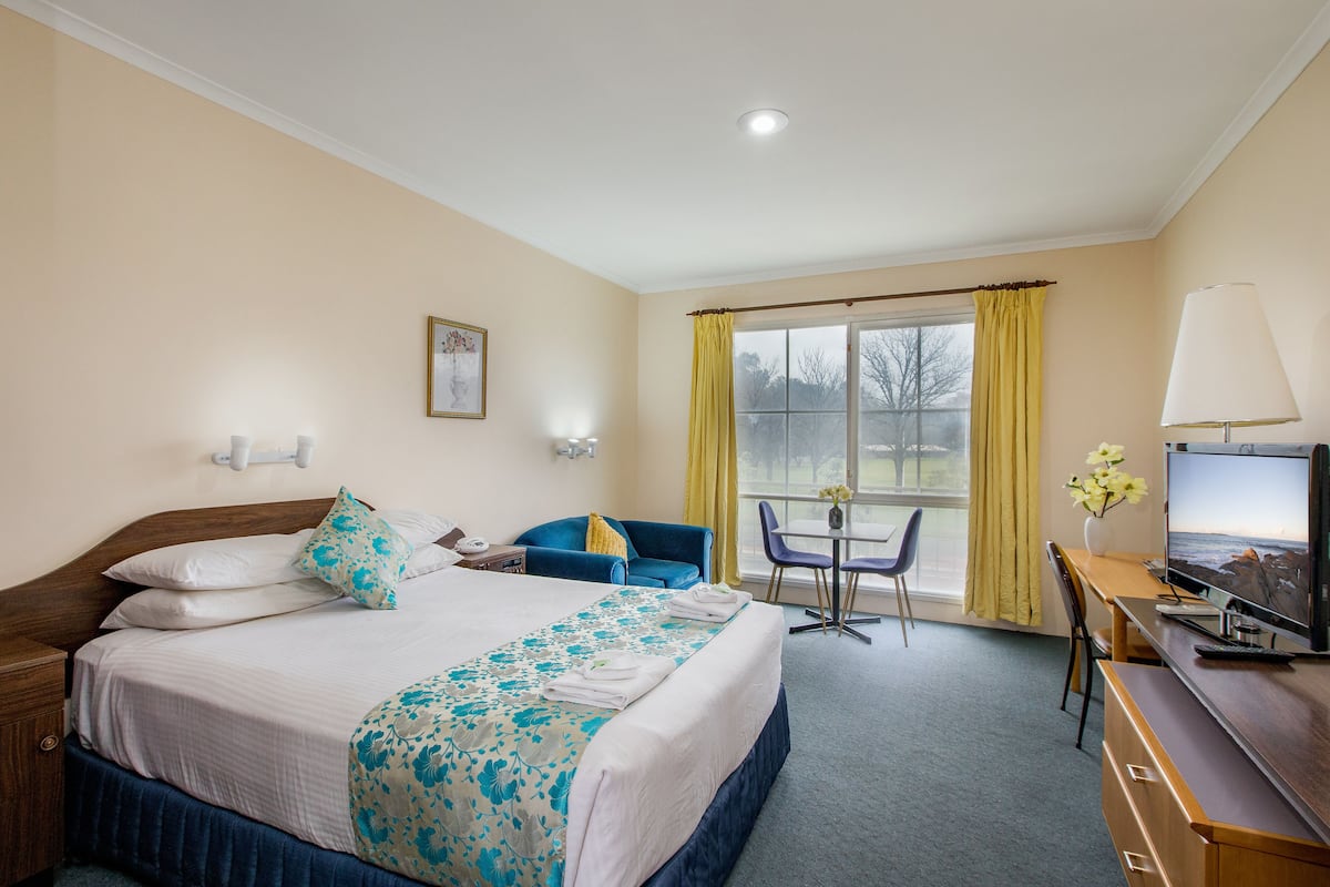 A hotel room with a bed, a desk with a computer, a chair, a window with curtains, and a lamp.