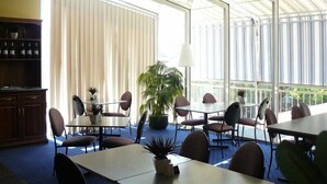Una habitación amplia con ventanas grandes, un área de comedor con mesas y sillas, y una planta en maceta.