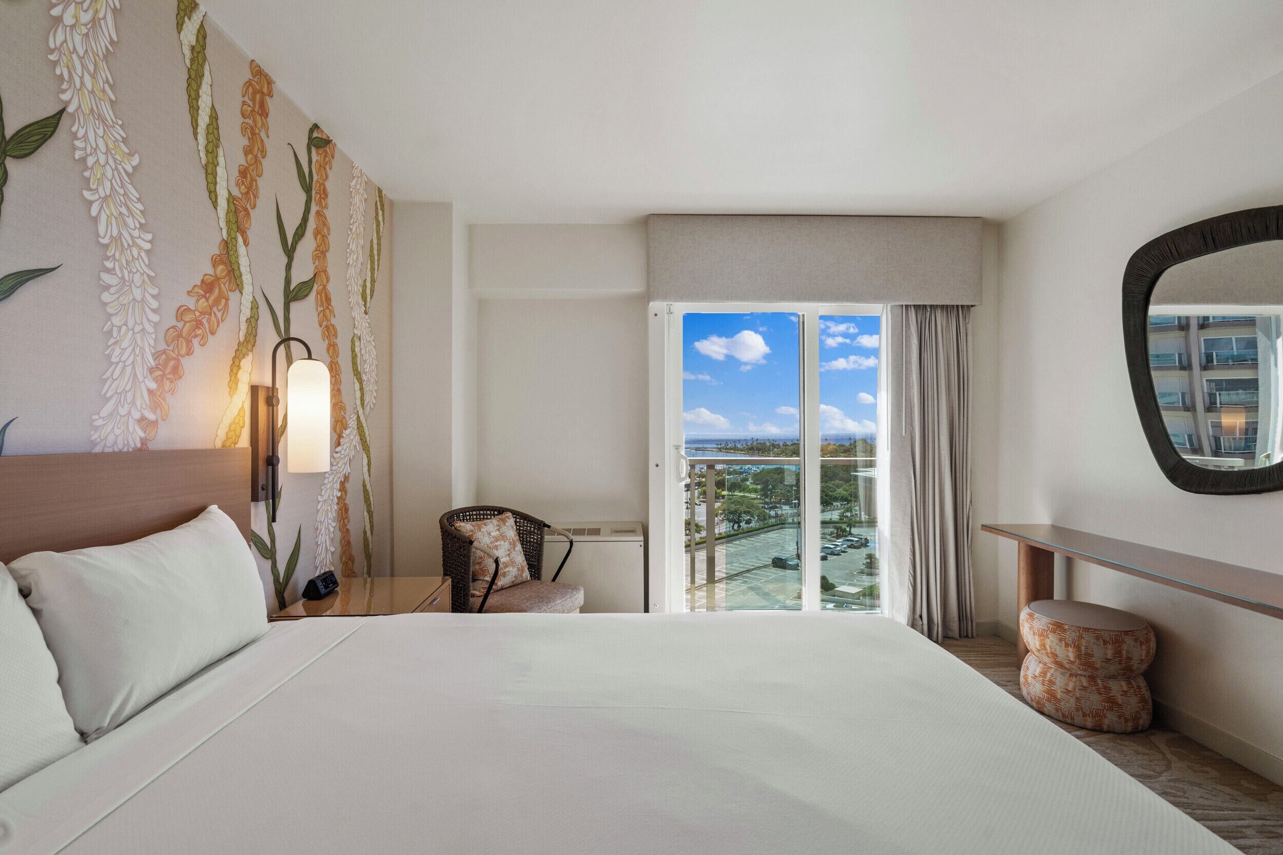 Habitación de hotel con una cama grande, una silla, una mesita de noche, una lámpara, un espejo y un balcón con vistas.