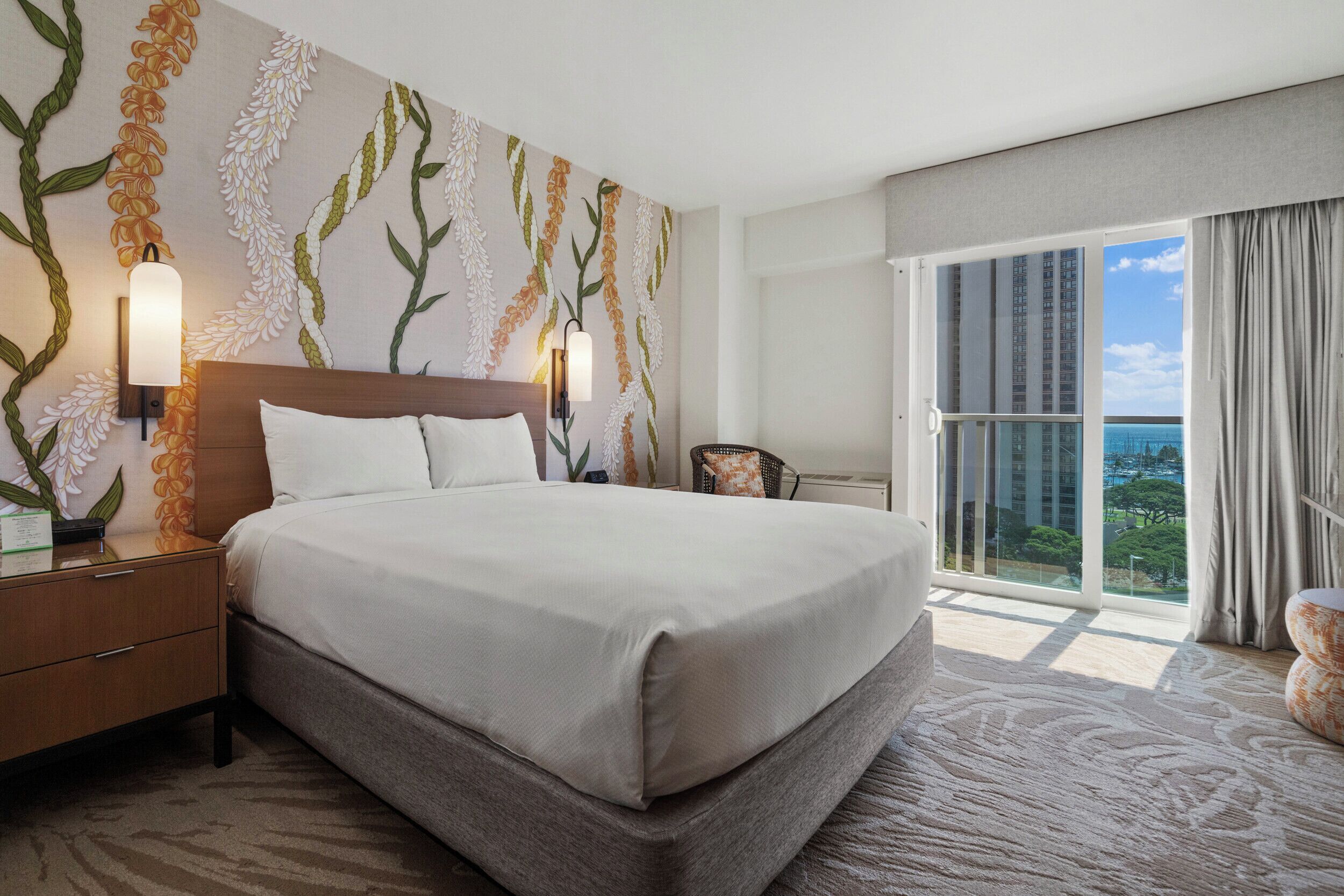 A hotel room with a large bed, a nightstand, a wall-mounted lamp, and a view of the ocean through a large window.