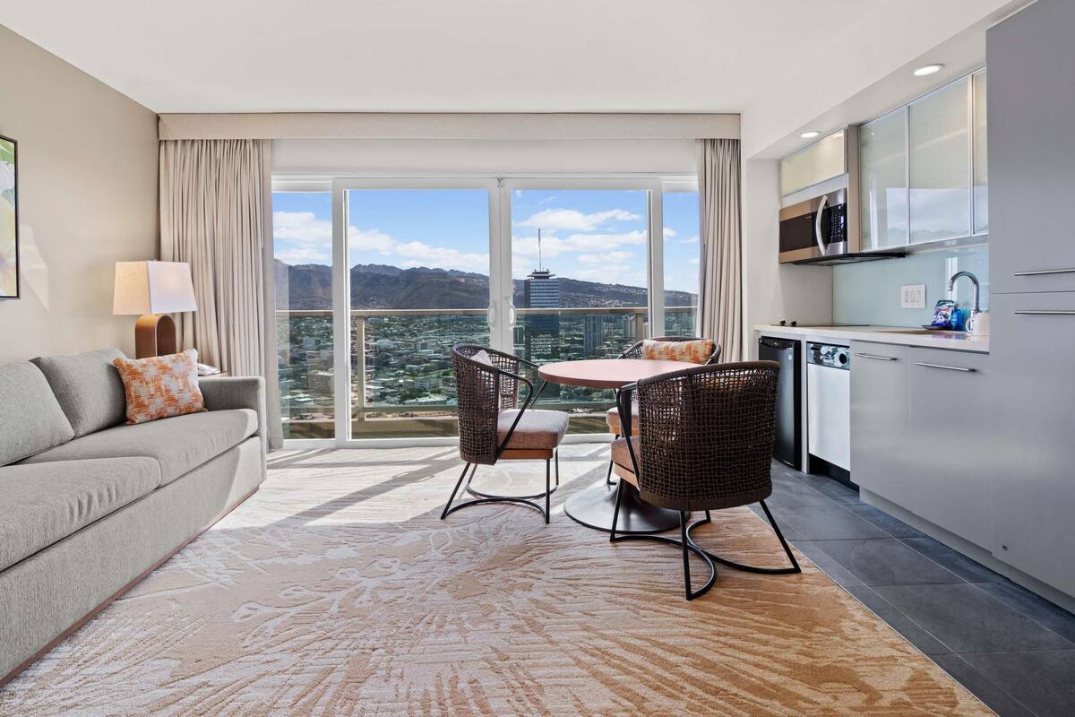 Una habitación de hotel moderna con balcón, un sofá, una mesa de comedor con sillas, una zona de cocina y un ventanal con vistas a la ciudad.
