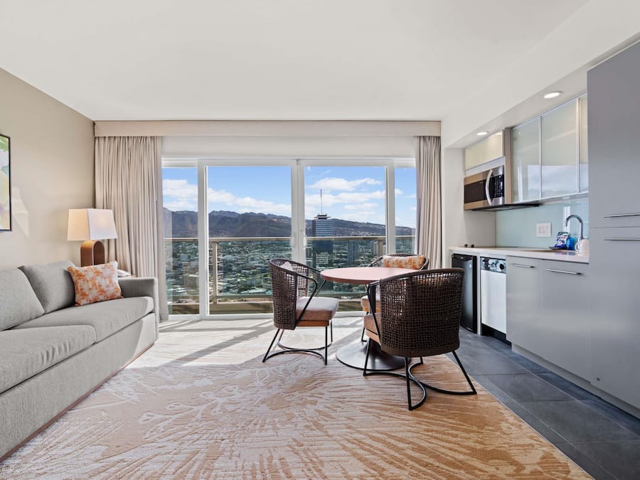 Una habitación de hotel moderna con balcón, un sofá, una mesa de comedor con sillas, una zona de cocina y un ventanal con vistas a la ciudad.