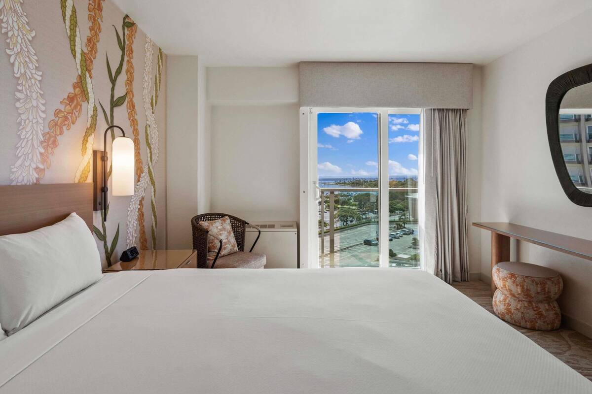 Habitación de hotel con cama, una silla, mesita de noche, lámpara, una mesa redonda y un ventanal con vistas.