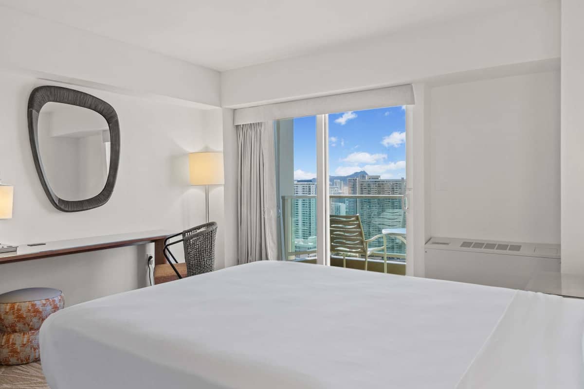 Habitación de hotel con una cama grande, un escritorio con lámpara, una silla, un otoman redondo, un espejo grande y vistas a edificios altos.