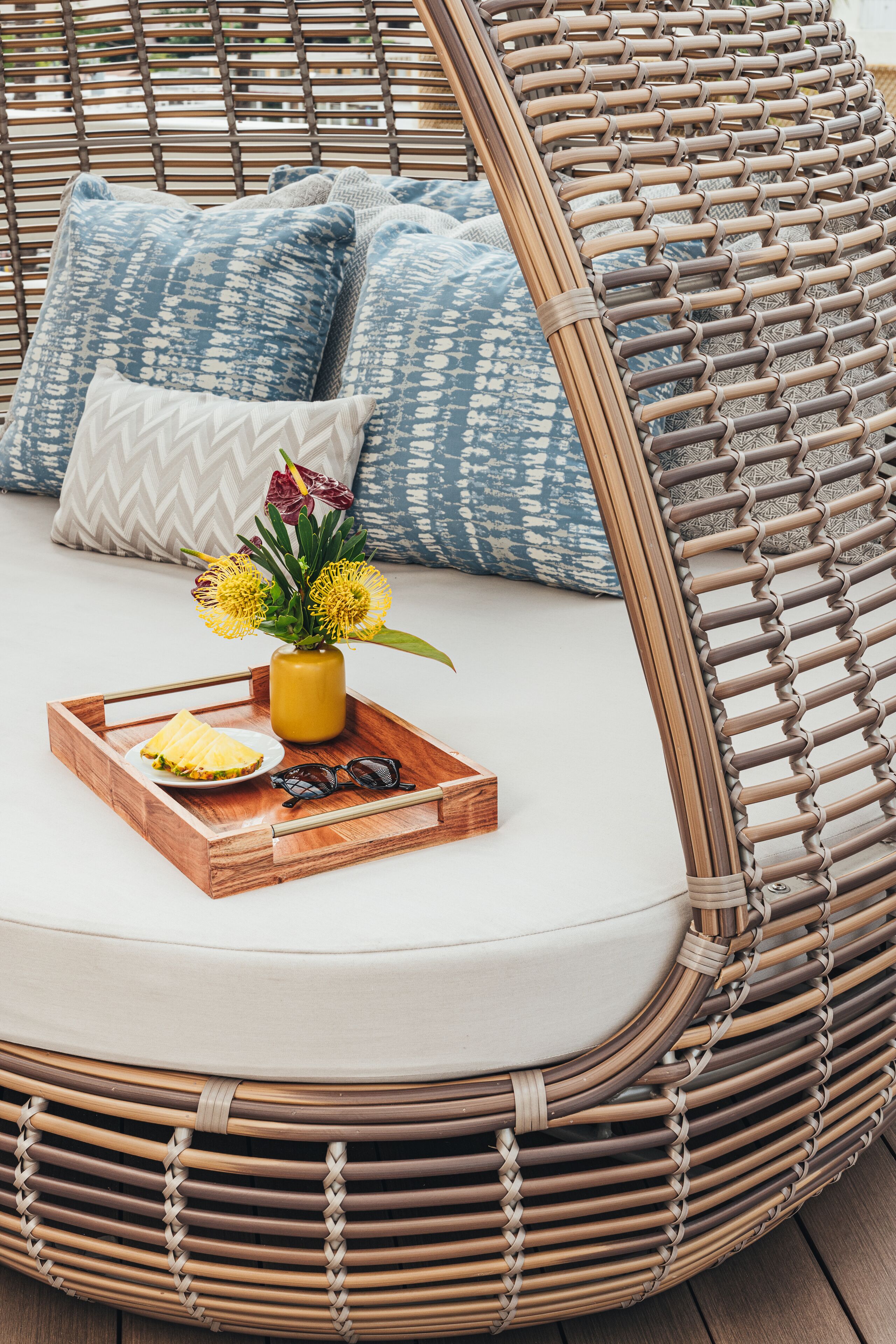 Un fauteuil de relaxation en osier, doté d’un siège et d’un dossier rembourrés, accompagné de coussins assortis et d’un plateau sur lequel se trouvent un vase de fleurs, des lunettes de soleil et un livre.
