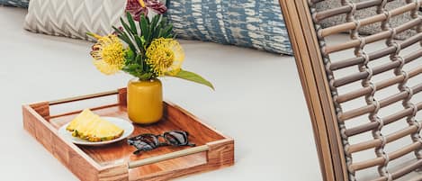 A wicker lounge chair with a cushioned seat and backrest, accompanied by matching pillows and a tray with a vase of flowers, sunglasses, and a book.