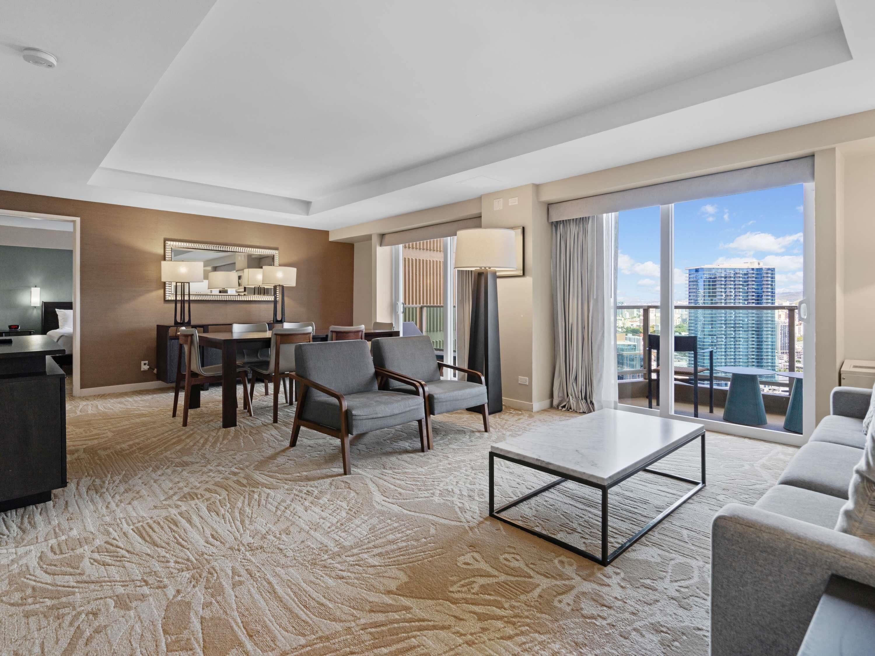 Habitación de hotel con comedor, sofá, mesa de centro y un amplio ventanal con vistas a la ciudad.