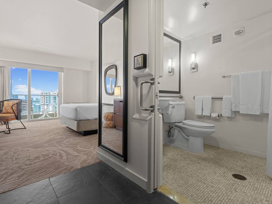 A modern hotel room with a large window, a balcony, a bathroom with a toilet and sink, and a seating area with a sofa and chair.