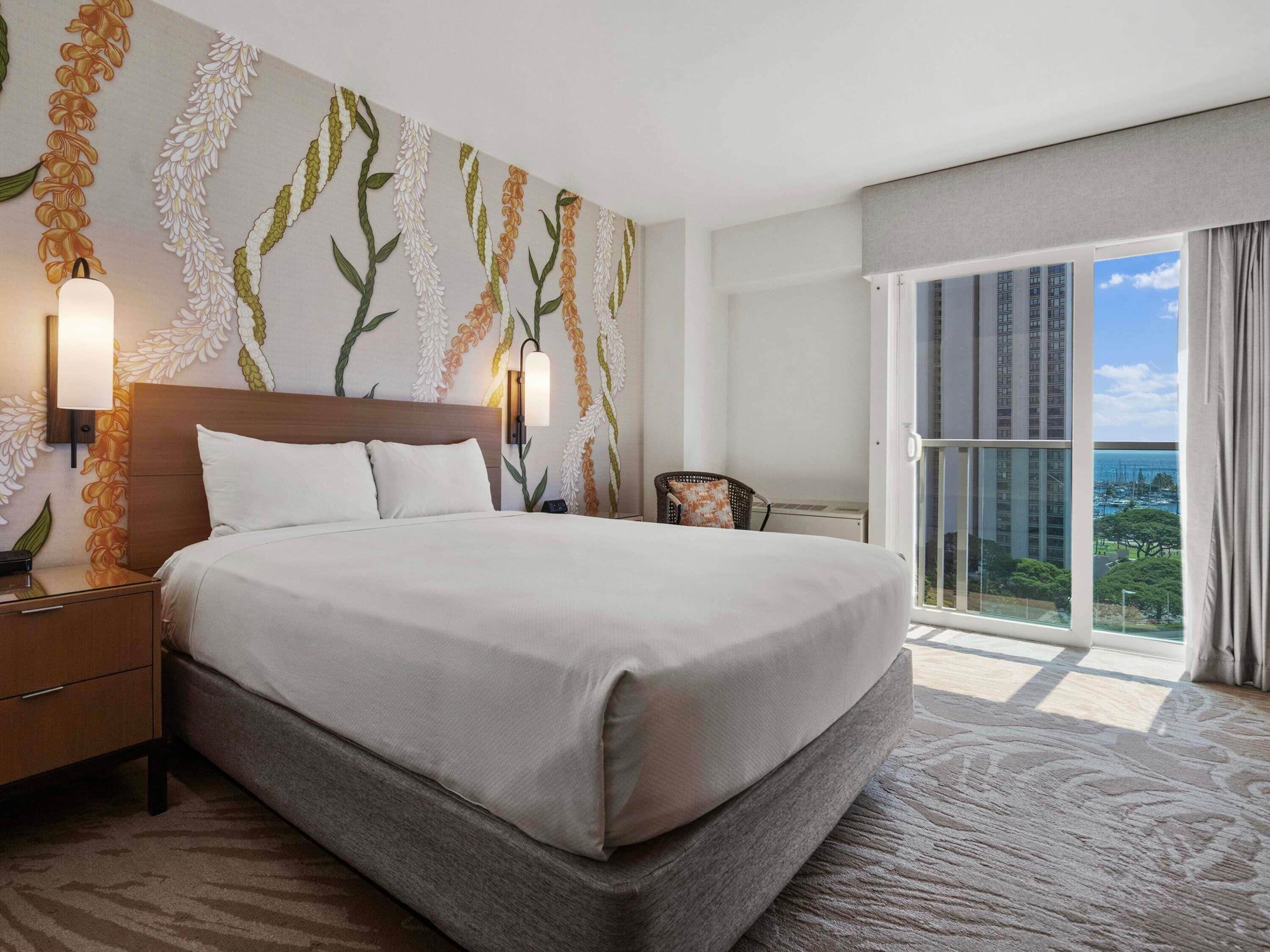A hotel room with a large bed, a nightstand, a wall-mounted lamp, a chair, and a view of the city through a large window.