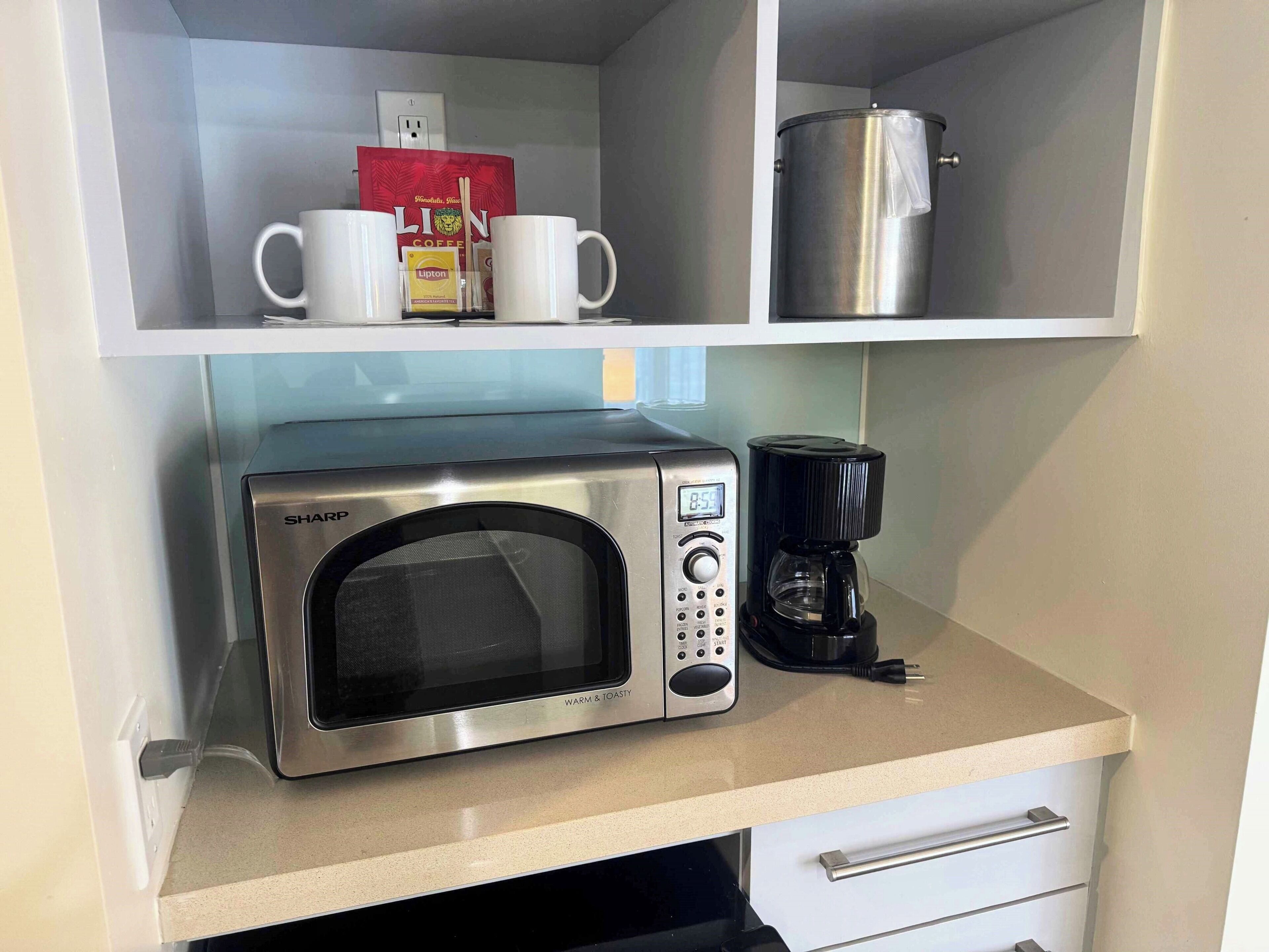 A microwave oven with a digital display, a coffee maker, and a set of white mugs on a kitchen counter.