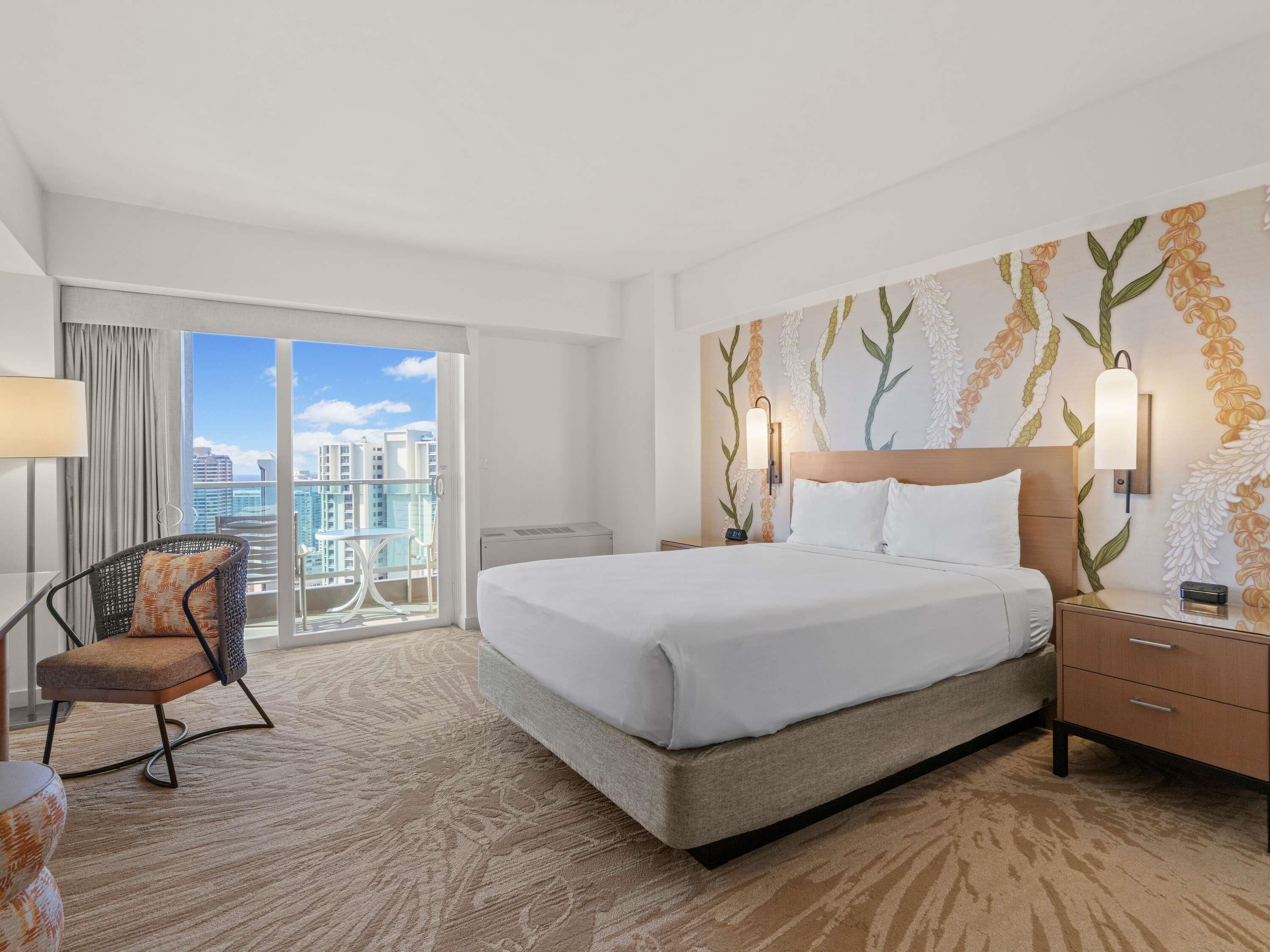 A hotel room with a large bed, a chair, a nightstand, and a view of the city through a balcony.