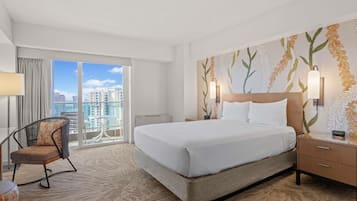 A hotel room with a large bed, a chair, a nightstand, and a view of the city through a balcony.