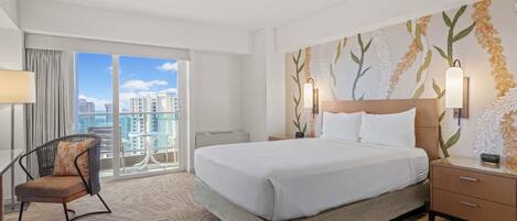 A hotel room with a large bed, a chair, a nightstand, and a view of the city through a balcony.